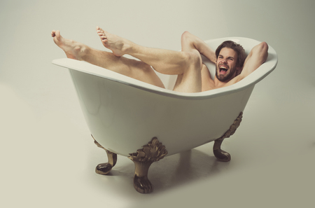 Angry man or unshaven caucasian macho with stylish haircut, hair, with naked, sexy muscular body, legs, feet, arms with biceps, triceps shouting in white, classic bath, tub on grey backgroundの写真素材