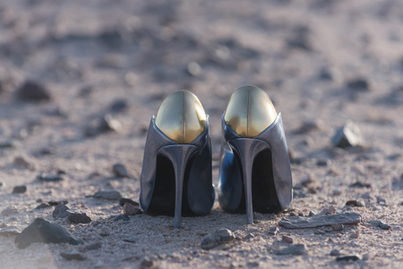 fashion and glamour. fashionable pair of female shoes, high heels, golden and silver, on ground or soil of sand and rocky stones, desert surface on sunny day outdoors on grey sandy background.の写真素材