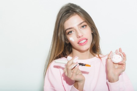 Pretty happy girl or beautiful woman with long, blond hair, putting facial cream or mask on smiling face skin with brush from jar on white background. Cosmetology, beauty and spa, copy spaceの写真素材
