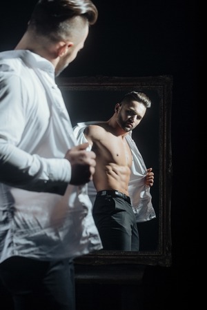handsome muscular macho man or athlete guy with sexy body of bodybuilder with torso, belly, six packs, has beard on serious face in pants, white shirt near vintage mirror isolated on black backgroundの写真素材