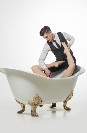 Handsome man or fashion model with stylish haircut, hair, relaxing with sexy, naked female legs, feet, in classic bath tub in fashionable wear, vest, shirt and pants isolated on white backgroundの写真素材