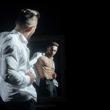 handsome muscular macho man or athlete guy with sexy body of bodybuilder with torso, belly, six packs, has beard on serious face in pants, white shirt near vintage mirror isolated on black backgroundの写真素材