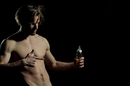 Handsome man, unshaven macho, bodybuilder, with stylish hair, sexy, naked, muscular torso with six packs, abs, biceps, triceps pointing at feeding baby bottle with teat on black background, copy spaceの写真素材