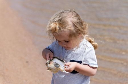 discovery and underwater. cute baby boy, small, little child with blond hair with seashell or marine shell on beach at sea shore with crystal clear water on sunny on sandy background. summer vacationの写真素材