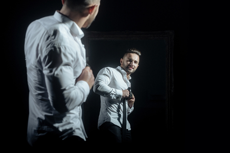 handsome muscular macho man or athlete guy with sexy body of bodybuilder has beard on happy face in white shirt, tie near vintage mirror isolated on black backgroundの写真素材