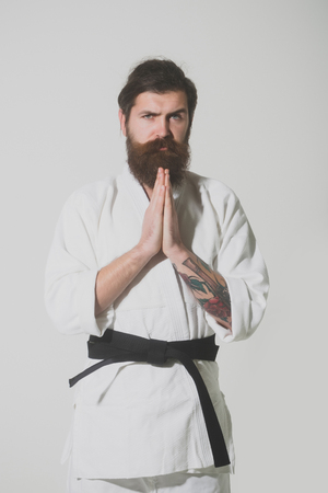 bearded karate man, long beard, brutal caucasian hipster with moustache in white kimono with black belt holds hands posing with serious face on grey background, unshaven guyの写真素材
