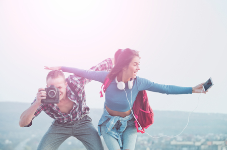 Smiling happy couple pretend flying in sky. Handsome man or photographer photographing with camera. Pretty girl or cute woman with smartphone, earphones and backpack. Traveling, vacationの写真素材