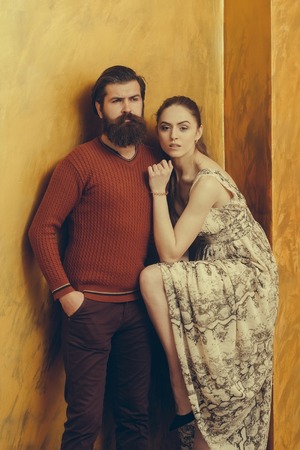 Serious bearded man with long beard, hipster, in sweater and pretty girl or beautiful woman in sexy dress with raised sexi leg in black high heel shoe on yellow wallの写真素材
