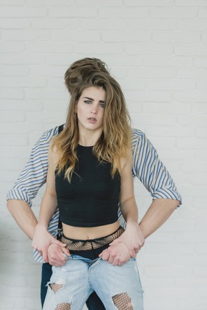 Stylish woman in sexy lingerie, black top, panties and fishnet tights, pantyhose. Handsome man undressing blue jeans from pretty girl on white wall. Seduction and passionの写真素材