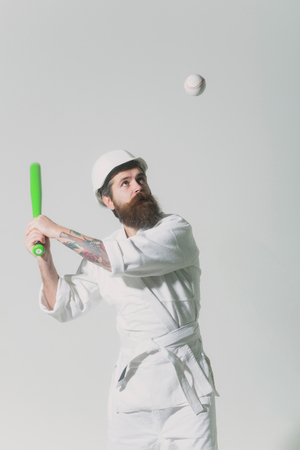 bearded karate man, long beard, brutal caucasian hipster with moustache in white kimono with black belt, builder helmet holds green baseball bat with ball on grey background, unshaven guyの写真素材