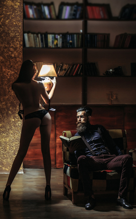 young couple of handsome bearded man sitting on armchair in library reading book and pretty cute girl or woman in black lingerie with sexy buttocks and body holding lampの写真素材