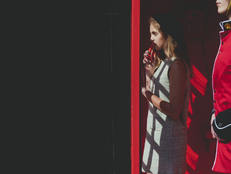 Pretty girl or young woman, teenager, talking on phone in retro, public, red telephone box, kiosk next to queens guard, soldier, on sunny day on black wall. traveling and destinations, copy spaceの写真素材