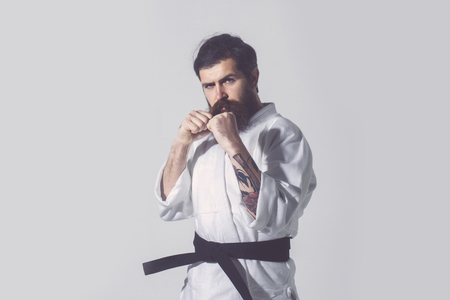 bearded karate man, long beard, brutal caucasian hipster with moustache in white kimono with black belt holds hands posing with serious face on grey background, unshaven guy, copy spaceの写真素材