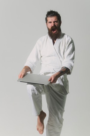 bearded karate man, long beard, brutal caucasian hipster with moustache in white kimono holds laptop with shouting face on grey background, unshaven guyの写真素材