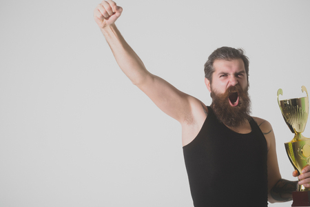 bearded man, long beard, brutal caucasian hipster with moustache in black vest holds gold champion cup with happy face on grey background, unshaven guy, copy spaceの写真素材