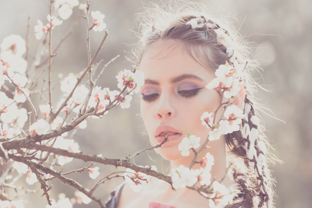 pretty girl or woman with fashionable makeup on cute face, has stylish hair, posing among blooming spring flowers or cherry blossom white color on tree branch sunny day outdoor on natural backgroundの写真素材