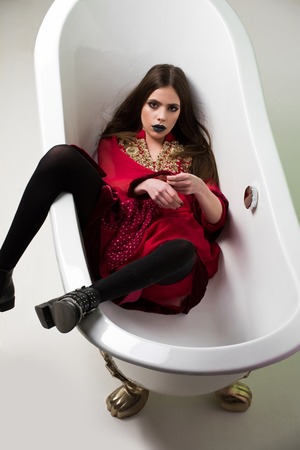 woman or pretty young caucasian brunette girl in red dress, boots with long hair and black lipstick on lips in bath tub on golden legs in bathroom in dress on white backgroundの写真素材
