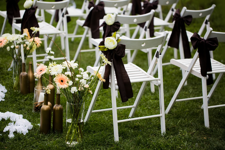 White wooden chairs decorated with black bows and roses, fresh, blossoming flowers, for holiday or anniversary ceremony in row outdoors on green grass background. Wedding and celebrationの写真素材