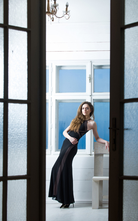Pretty girl or beautiful fashion woman with long blond hair in black dress holding brown cup seen through open doors on window backgroundの写真素材