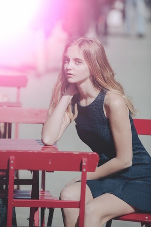 Summer and relaxation, Pretty girl or young woman, teenager, with cute young face and stylish, blond, long hair in fashion dress waiting for order at red table in street cafe in sunlight on summer dayの写真素材