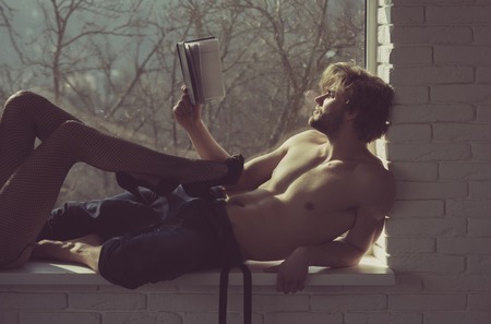 Muscular man or macho with fit, naked torso and female legs in erotic, black, fishnet tights and shoes with high heels on window sill on blurred natural background. Reading book and relaxの写真素材