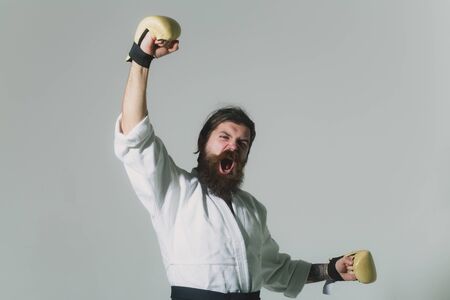 bearded karate man, long beard, brutal caucasian hipster with moustache in white kimono with black belt and boxing gloves with shouting happy face on grey background, unshaven guy, copy spaceの写真素材