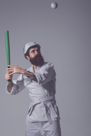 bearded karate man, long beard, brutal caucasian hipster with moustache in white kimono with black belt, builder helmet holds green baseball bat with ball on grey background, unshaven guyの写真素材