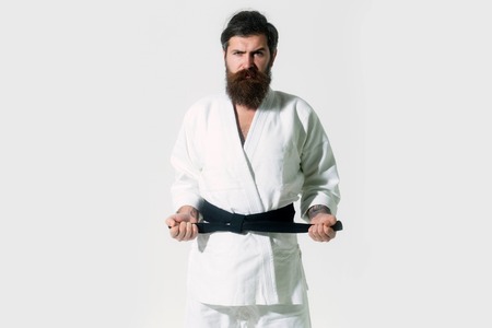 bearded karate man, long beard, brutal caucasian hipster with moustache in white kimono with black belt holds hands posing with serious face on grey background, unshaven guy, copy spaceの写真素材