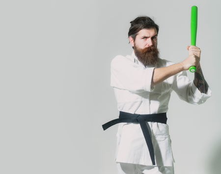 bearded karate man, long beard, brutal caucasian hipster with moustache in white kimono with black belt holds green baseball bat with serious face on grey background, unshaven guy, copy spaceの写真素材