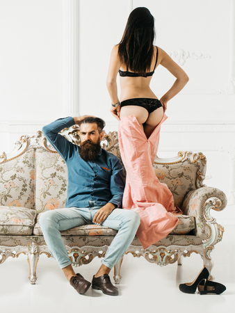 young couple of handsome bearded man in blue shirt and jeans with pretty cute girl or woman in black panties topless with sexy buttocks on luxurious vintage couch or sofa near shoes on whiteの写真素材