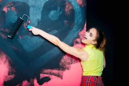 pretty happy woman builder or cute sexy girl painter with roller, building tool painting pink wall in black, has brunette hair in yellow vest and red checkered skirt. repair and construction conceptの写真素材