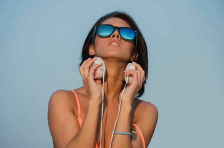 Pretty girl or sexy woman with brunette hair, looking at blue sky in fashion sunglasses and peach color shirt with headphones on sunny day. Summer vacation and leisureの写真素材