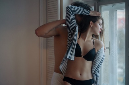desire and temptation, young sexy couple embrace near window, pretty girl with long hair in erotic lingerie and shirt on slim body and handsome man, love and relations, passion and tendernessの写真素材