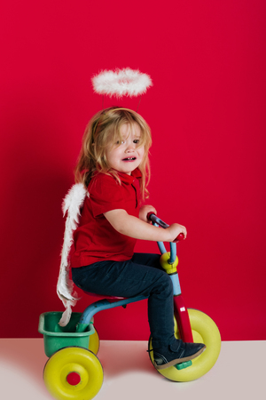 crying little boy with angel wings and halo has long blonde hair on bike on red backgroundの写真素材