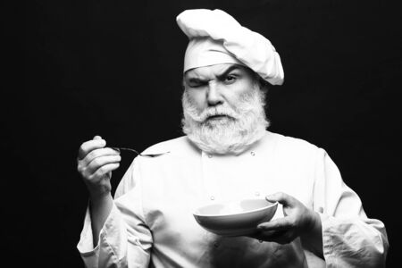 Bearded man cook in hat tasting food with spoon in studio, black and whiteの写真素材