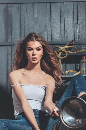 pretty girl or sexy woman biker with curly, long, blond hair in erotic undershirt sitting on vintage motorcycle, bike, in garage on grey wooden background. Motorcycling, hobby and lifestyleの写真素材