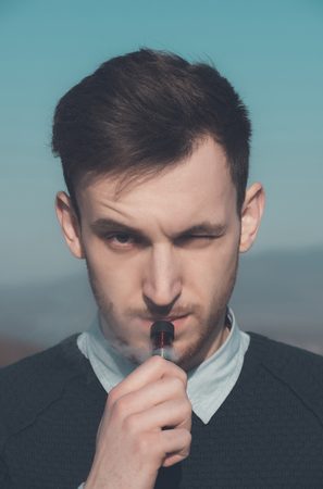 Unshaven, handsome, young man smoking or vaping electronic cigarette, with vapor cloud and squinting...