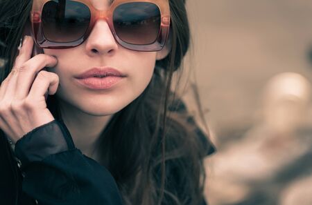 pretty woman, brunette, in stylish, fashionable, vintage brown sunglasses and black coat posing outdoors on blurred natural background. Retro. Fashion and styleの写真素材