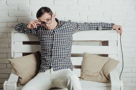 man in glasses with microphone, guy in checkered shirt and pants sit on wooden bench on white brick wall background with professional micの写真素材