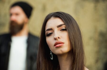 Pretty girl or cute woman with fashionable makeup on young face and stylish, brunette, long hair posing with blurred, bearded man with beard in background on grungy, cement wall. Beauty and fashionの写真素材