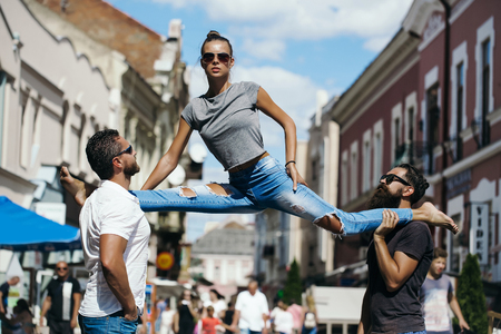 girl or pretty woman, fit model, in stylish sunglasses and blue jeans sitting leg split on shoulders of two bearded, handsome men on sunny, summer day on city street. Active lifestyle, friendsの写真素材