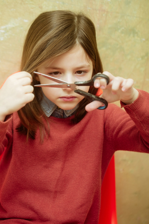 girl, small, little child or hairdresser, in orange sweater cutting long, brunette, hair with metallic scissors on beige background. Hairdressing and hairstyleの写真素材