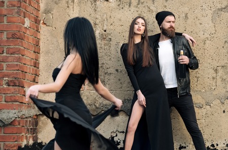 Young people or stylish friends having party on grungy cement wall. Two sexy girls or pretty women with long hair in black dresses and handsome, bearded man drinking wine from bottle. Lifestyleの写真素材