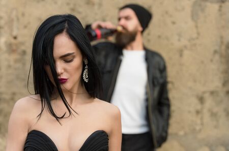 Unhappy sexy girl or pretty woman with beautiful makeup, stylish, long hair, black dress and blurred, brutal man with beard drinking wine in background on grungy, cement wall. Alcohol abuseの写真素材