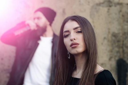 Adorable female with stylish, brunette, long hair and fashionable makeup on young face posing with blurred, bearded man with beard in background on grungy, cement wall. Beauty and fashionの写真素材