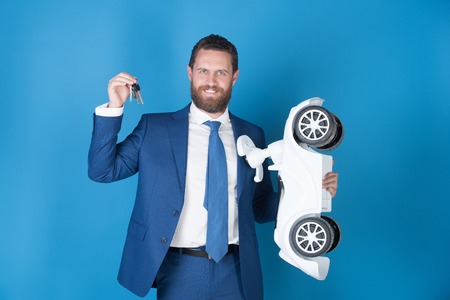 white toy car and keys in the hand of a happy smiling business man wearing suit on a blue backgroundの写真素材