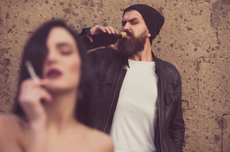 bad habits. Bearded brutal man or caucasian hipster with beard in leather jacket drinking wine from bottle on grungy cement wall. Blurred sexy girl or cute woman smoking cigarette in foreground. Unhealthy habitsの写真素材