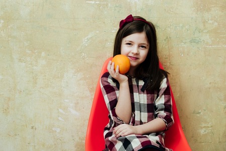vegetarian. Cheerful small, little girl, child with orange fruit in hand smiling in plastic chair on...