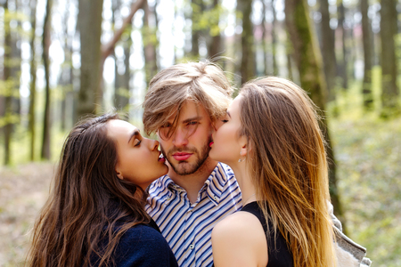 man getting kiss by two pretty girls or cute women on sunny day on natural background. Love triangle. Relationshipの写真素材