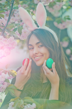 woman smiling with colored eggs, green and red, rosy bunny ears and long, brunette hair at tree with...